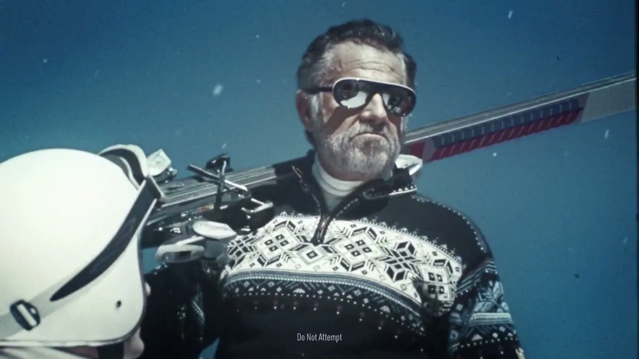 Jonathan Goldsmith in a patterned ski sweater and sunglasses, skis over one shoulder, standing beside a helicopter as a caption reads Do Not Attempt
