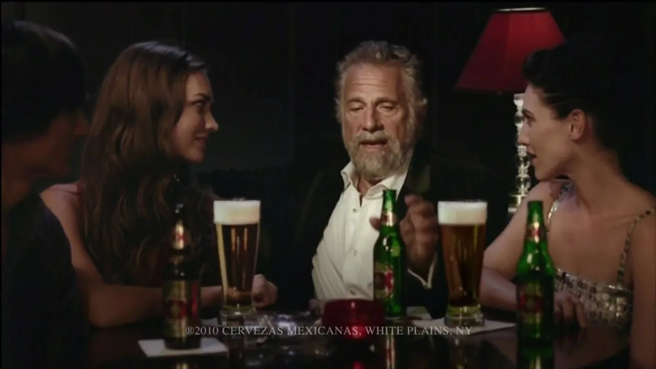 Goldsmith seated at a dark bar table between two women, several bottles and full glasses of Dos Equis in front of them